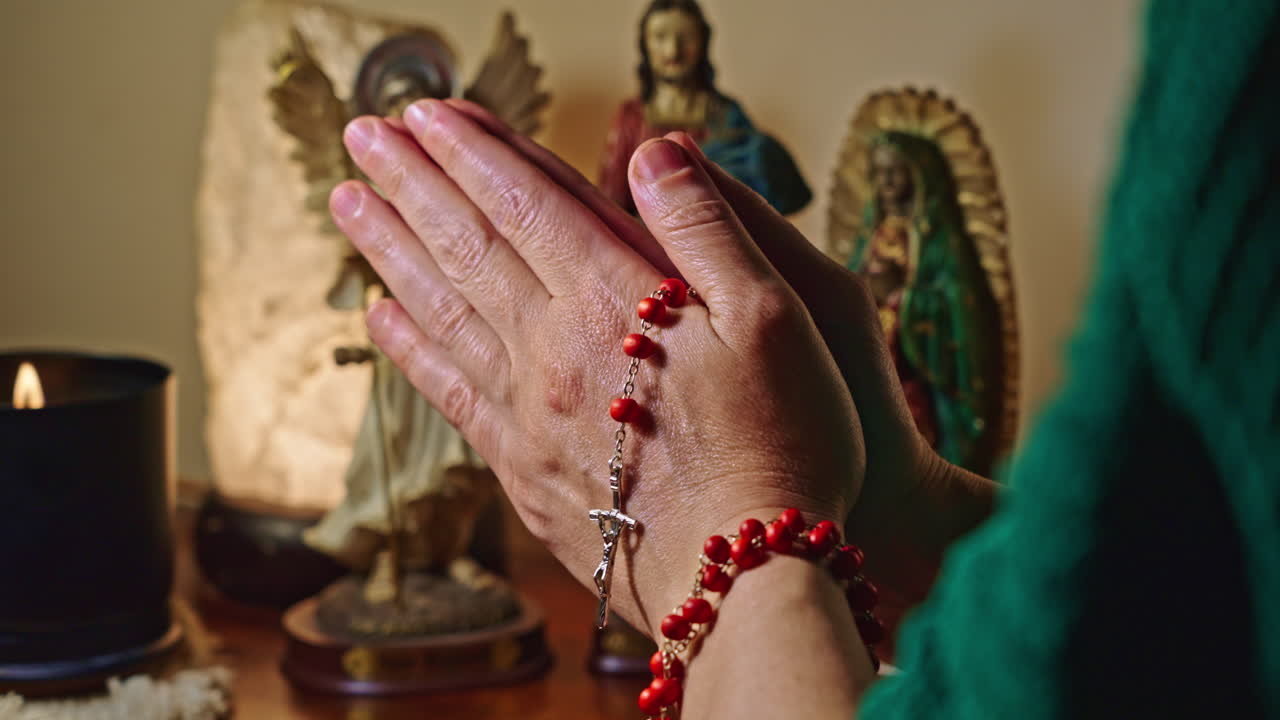 Primo piano di mani in preghiera con rosario e statue religiose.