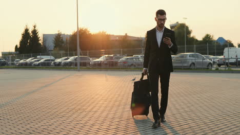 Handsome-Stylish-Man-In-Business-Outfit-With-A-Suitcase-On-Wheels-Walking-Next-To-The-Parking-Place-And-Tapping-While-Chatting-On-The-Smartphone