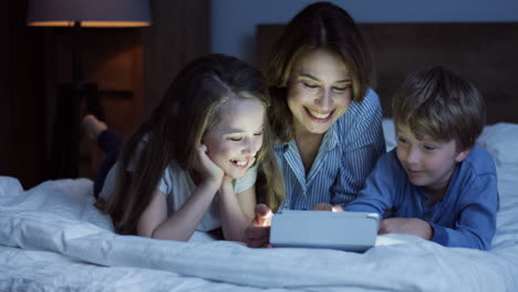 caucasian mother and his little son and daughter lying on the bed and watching something on the tablet