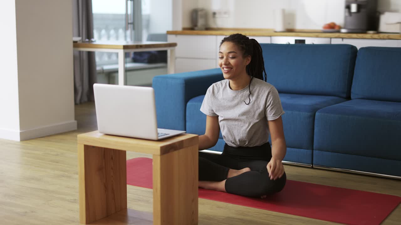 Smiling african american woman talking on video call on laptop while sitting on yoga mat