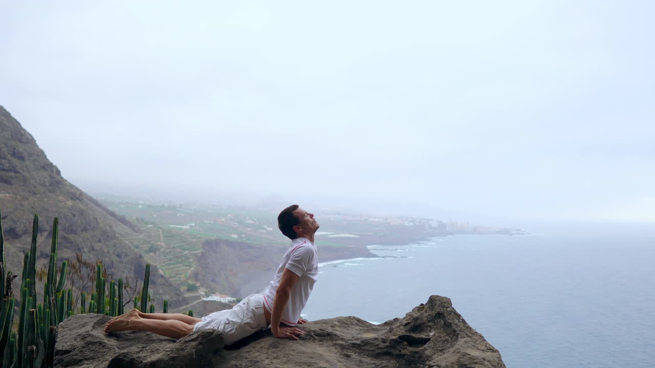 Positioned at a cliff's edge, the man assumes a dog pose, overlooking the ocean, inhaling the sea air as he embarks on a yoga journey through the islands