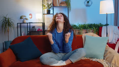 happy woman celebrating success showing thumbs up dancing sits on sofa in living room at home