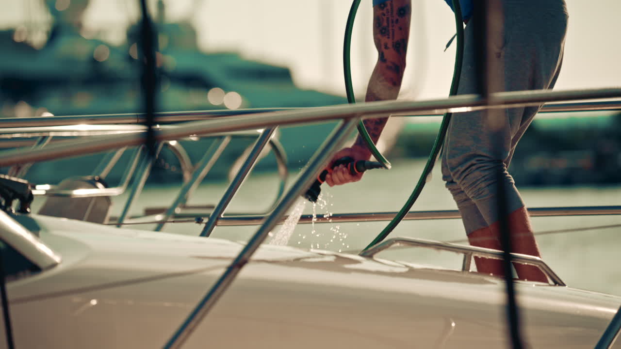 Hands washing a yacht's deck with a hose, with water spraying in the air and sunlight creating sparkling highlights on the boat's railings
