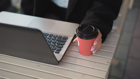 close-up of laptop keyboard with partial seating view, person with polished nails holding a tea cup next to laptop, dropping it onto table and beginning to type, fingers adorned with rings