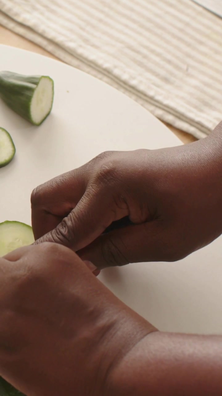 Video verticale: Mani adulte che prendono un coltello blu e affettano un cetriolo a rondelle su un tagliere per l'insalata.