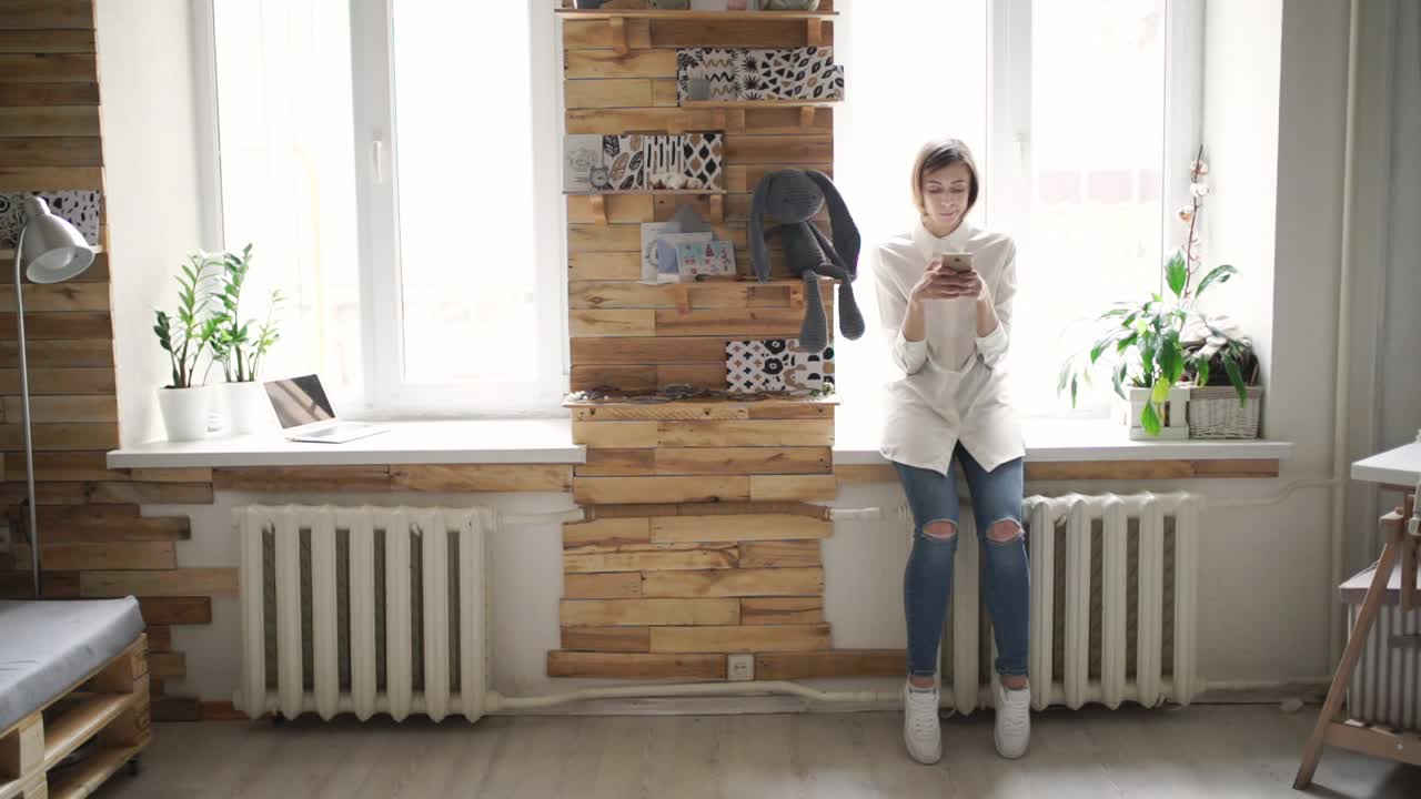 mujer joven con teléfono móvil en las manos y de pie en la ventana delantera