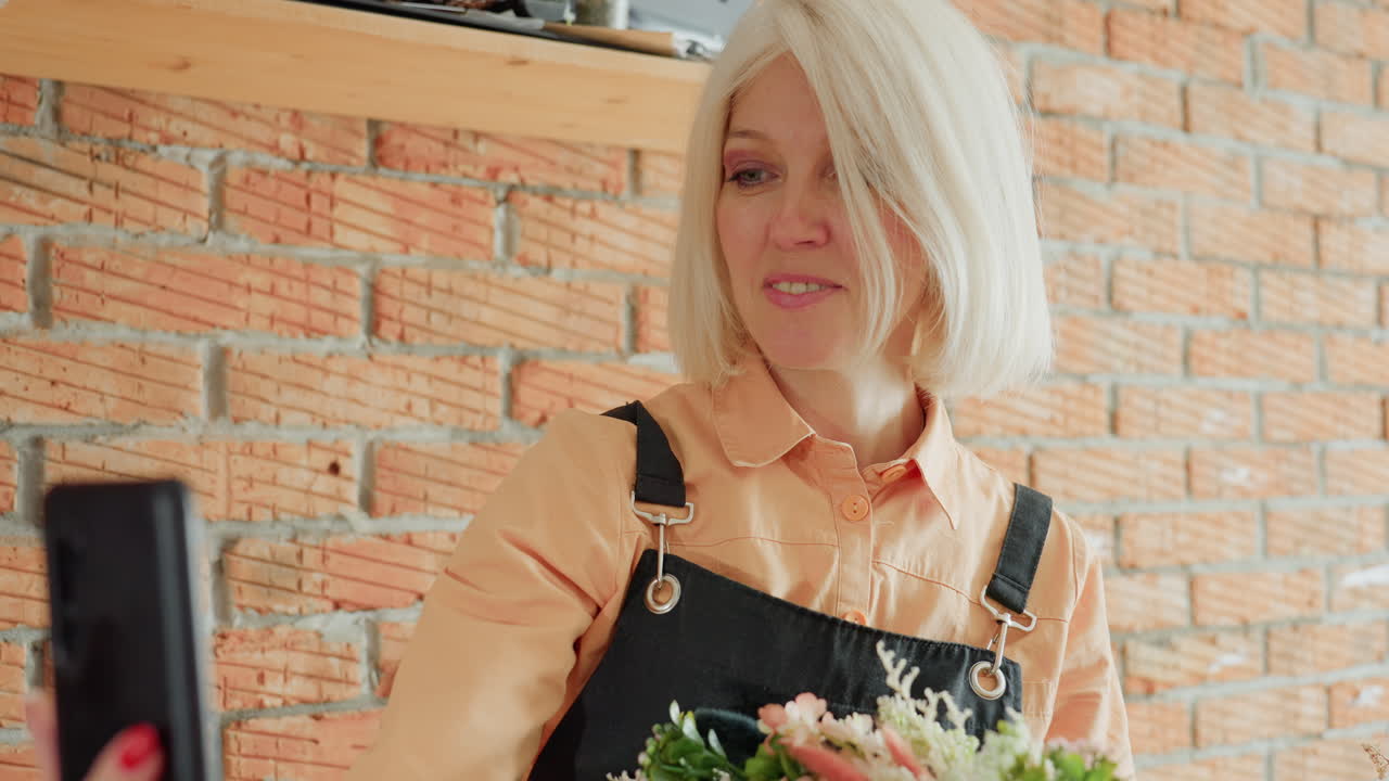 Smiling female florist in black apron holding floral wreath decorated with pink blossoms and greenery, taking selfie with smartphone in rustic workshop with brick wall background, joyful presentation