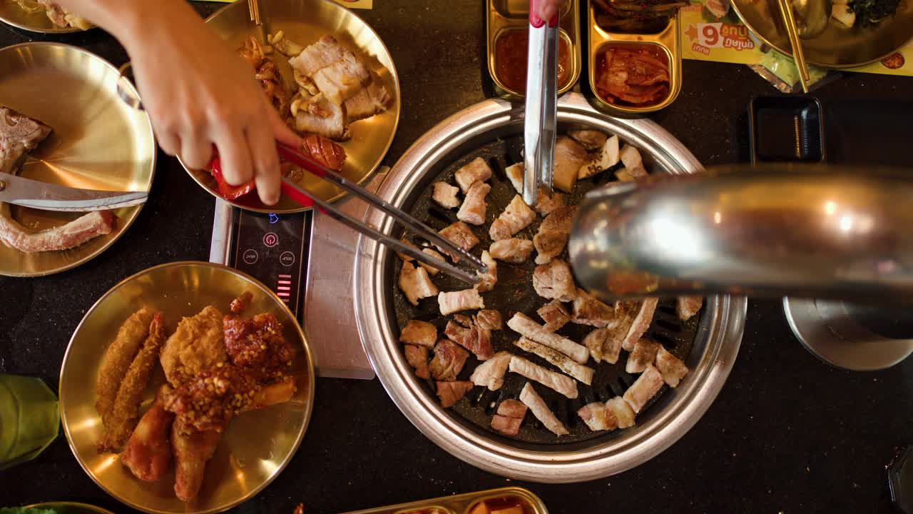 Hand uses tongs to turn pork belly on tabletop grill, warm lighting, overhead camera angle