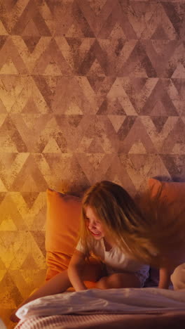 joyful little girl drops down pillow fighting with long haired mother on comfortable bed near play tent with glowing night lamp inside slow motion