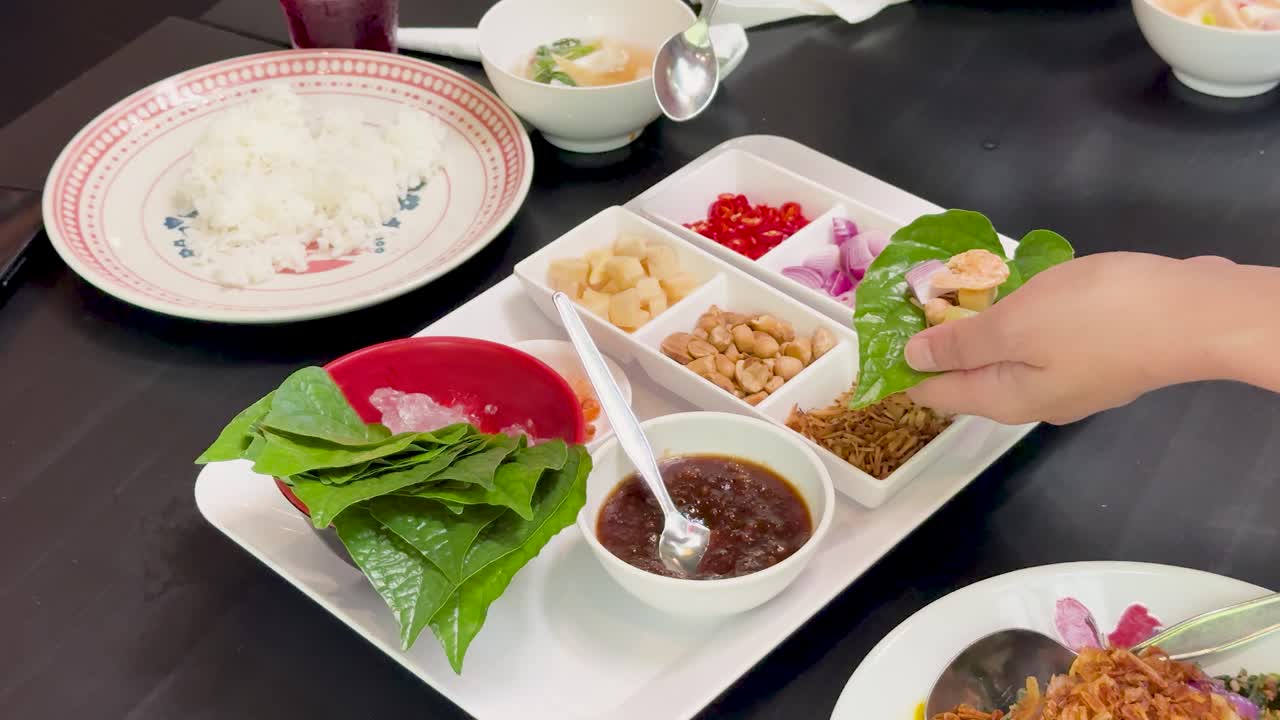 Hands prepare Miang Kham by adding various fillings and sauce to wild betel leaves on a neatly arranged tray, under bright indoor lighting