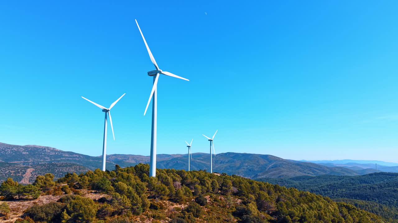 Alicante, Spagna, 8 agosto 2025: Turbine eoliche e luna su un crinale spagnolo. Turbine eoliche bianche su un crinale montuoso spagnolo con pineta, colline distanti e una piccola luna