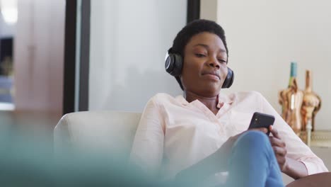 Feliz-Mujer-Afroamericana-Sentada-En-Un-Sillón-En-La-Sala-De-Estar,-Usando-Un-Teléfono-Inteligente