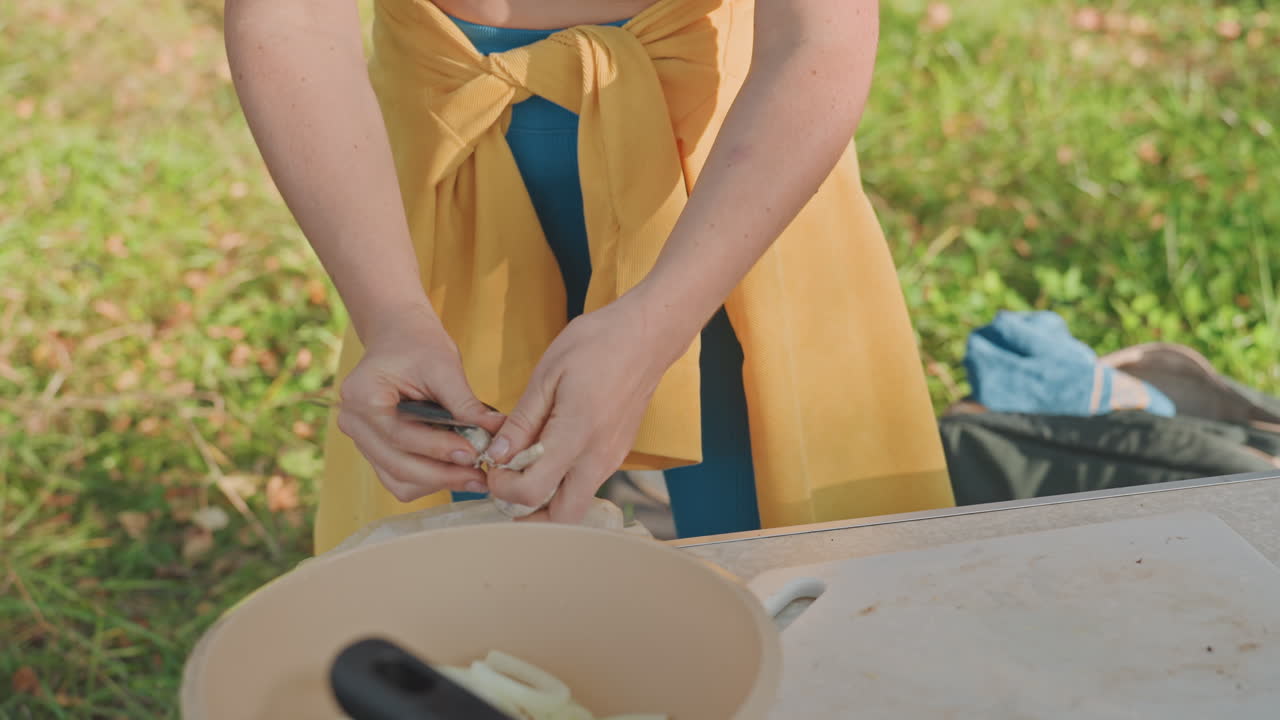 Wanita mengiris bawang di luar ruangan menggunakan wajan kemah, tangan yang stabil membimbing pisau di atas area memotong, meja piknik dengan mangkuk dan panci, antisipasi hidangan kemah yang mendesis, latar rumput cerah, persiapan yang efisien.