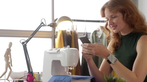 Female-Owner-Of-Fashion-Business-Using-Mobile-Phone-At-Sewing-Machine-In-Studio