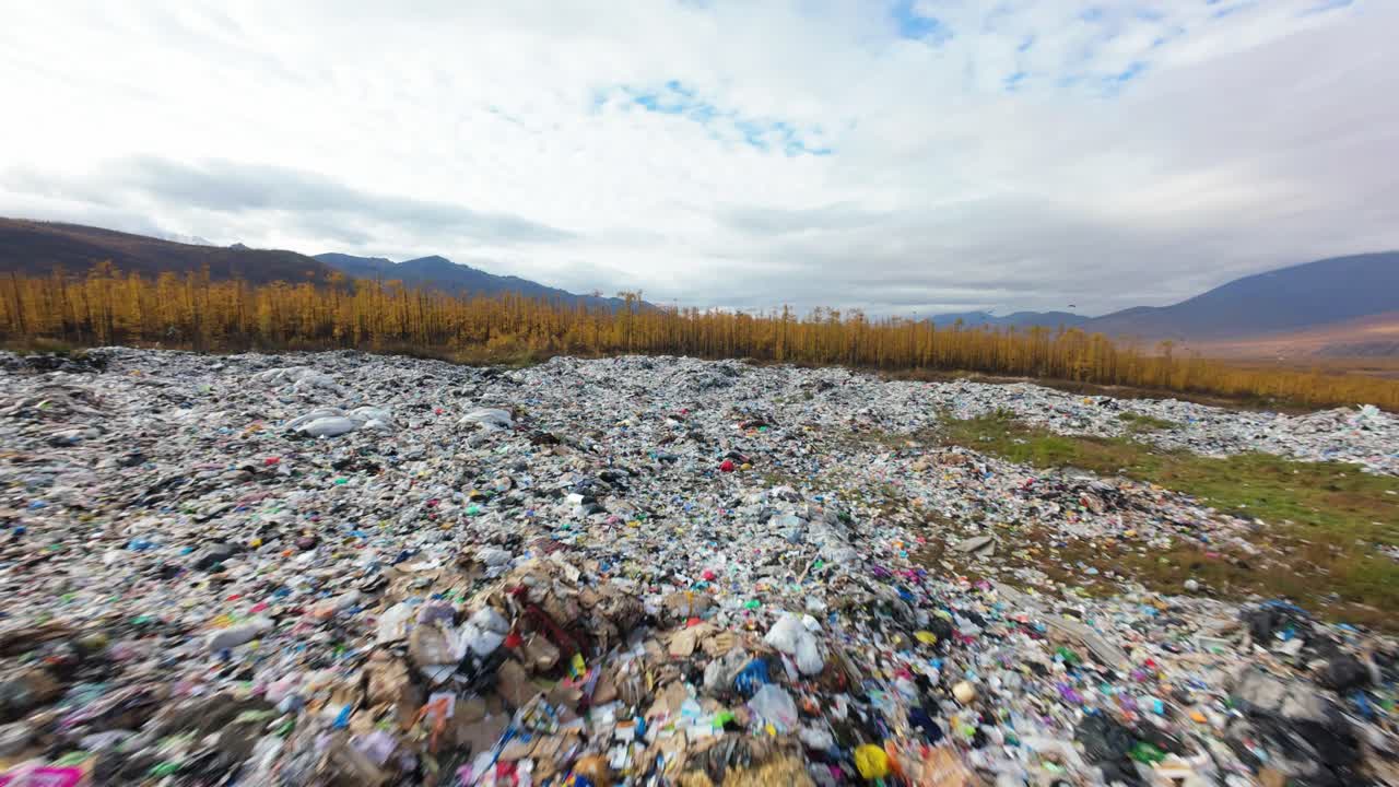 In a desolate landscape, remnants of consumer culture blend with nature, showcasing an extensive household waste dump surrounded by mountains