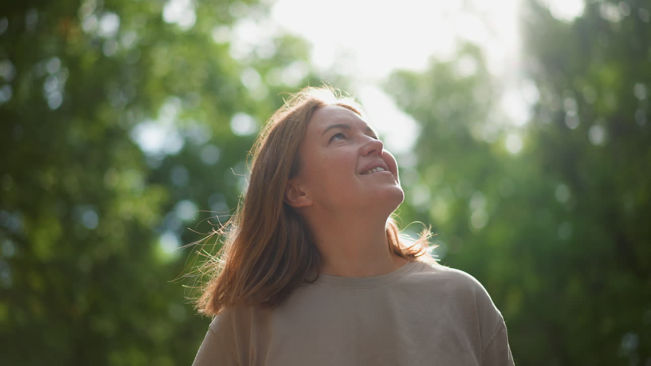 Donna bianca sorridente sotto alberi illuminati dal sole, profilo rilassato mentre la brezza le solleva i capelli durante una piacevole passeggiata nel parco. Il ritratto ambientale spontaneo evoca gioia, libertà e benessere consapevole, bokeh illuminato dal sole e vegetazione lussureggiante