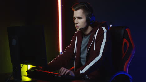 close up view of young serious man with headphones and playing a game on the computer in a room with colorful neon lamps on the wall 4