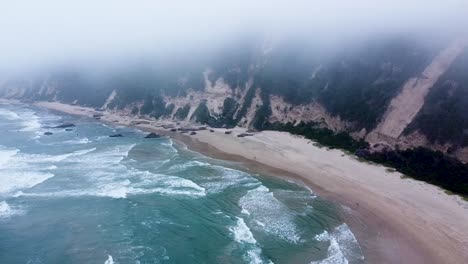 Tiro-Aéreo-De-Un-Dron-Que-Desciende-Debajo-De-Un-Manto-De-Nubes-Que-Se-Desplaza-Para-Revelar-Una-Playa-Desierta-En-Un-Día-Miserable,-Sedgefield,-Cabo-Occidental,-Sudáfrica