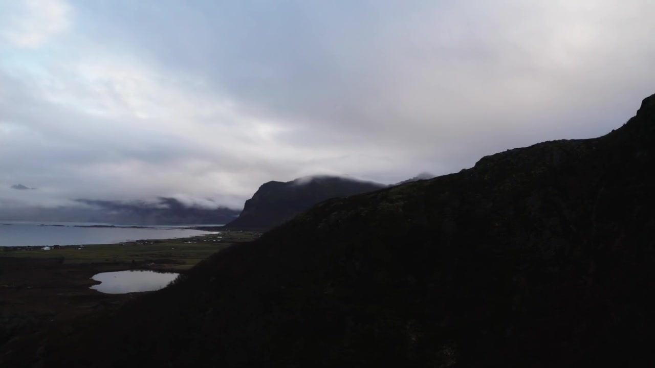 Tracking wide aerial shot of a mountain ridge leading to a lake and distant mist-covered mountain ranges under dark clouds, separated by a barren valley, creating a dramatic wilderness scene.