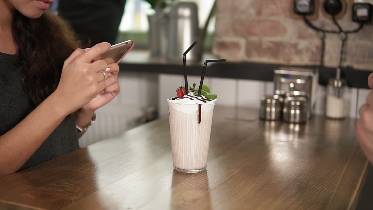 Hands of man and woman taking pictures of a glass with milk shake with cherry on the top and straw on a wooden table in cafe