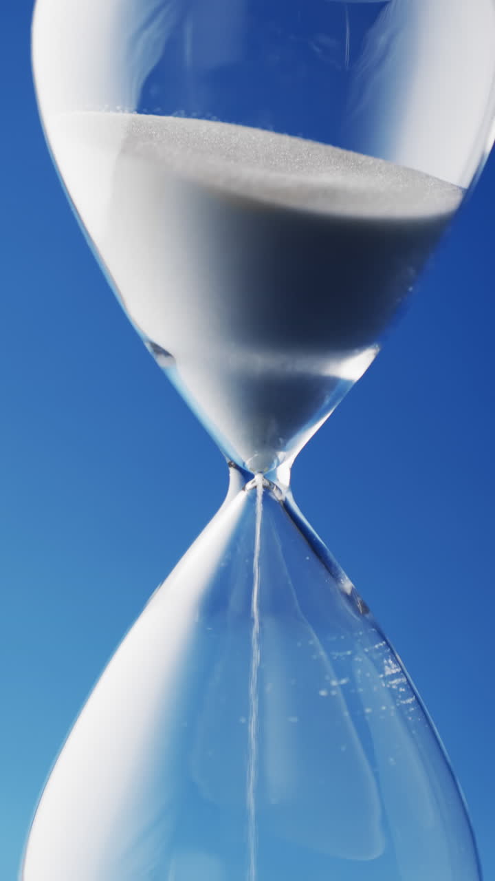 Vertical video of close up of hourglass with white sand pouring with copy space on blue background