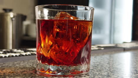 orange beverage with ice cube in old-fashioned glass. a slice of lemon is put into the drink with tongs. close up. blurred backdrop.