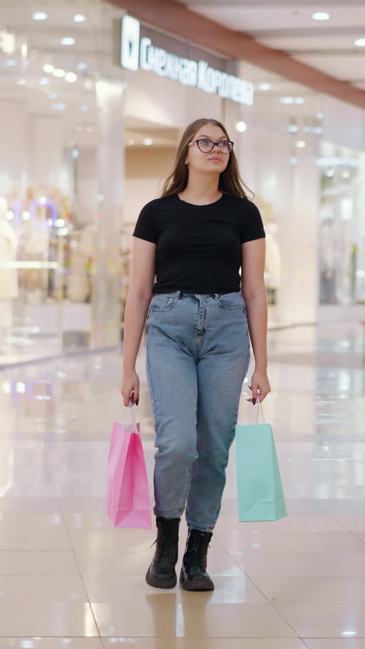 mujer en vaqueros y camiseta negra llevando bolsas de compras, caminando en un centro comercial moderno y bien iluminado con otros compradores en el fondo y la luz reflejada en las baldosas