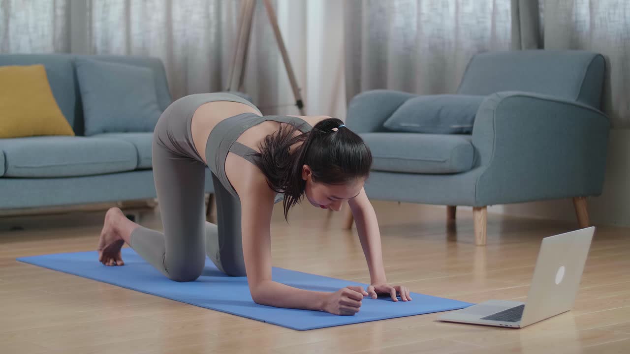 joven atleta asiática en ropa deportiva haciendo yoga en el antebrazo en la postura de tabla en la alfombra en casa con el entrenador en línea a través de una computadora portátil