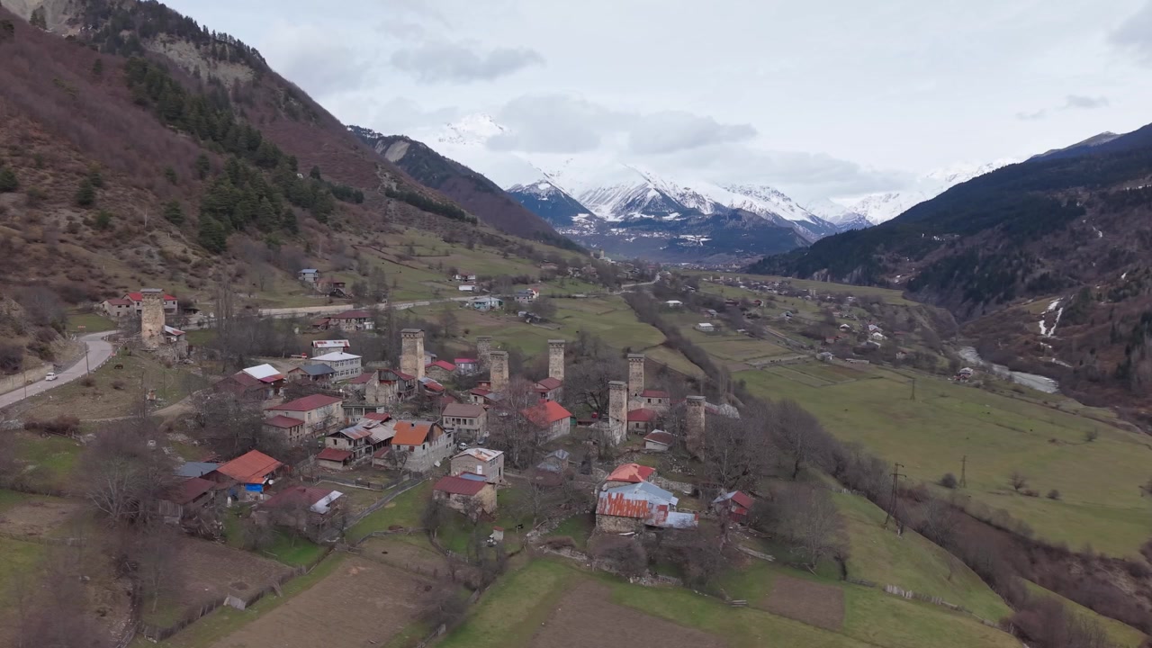 Orbita di drone che mostra le torri Svan medievali e le case tradizionali di Mestia, incorniciate da vette innevate e dal colorato paesaggio di inizio primavera della Georgia