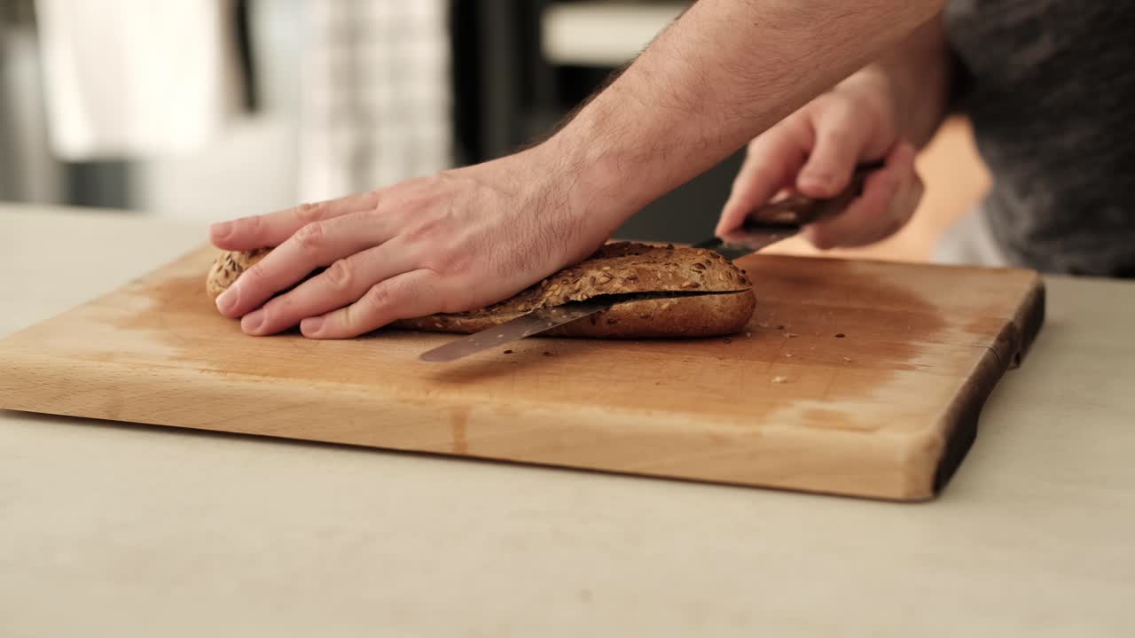 White man slicing through brown baguette with serrated knife on kitchen countertop