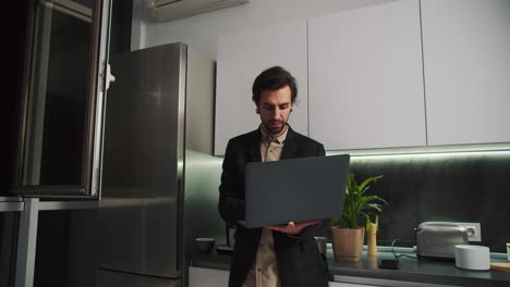 A-serious-brunette-man-with-stubble-in-a-black-jacket-and-beige-shirt-stands-holding-in-his-hands-a-gray-laptop-working-on-it-only-in-beige-shorts-in-a-modern-kitchen-in-an-apartment-while-working-remotely-in-the-evening