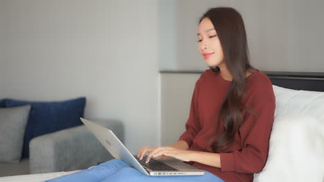 Young-woman-using-a-laptop-on-her-knees-while-sitting-on-a-bed-and-typing-on-the-keyboard