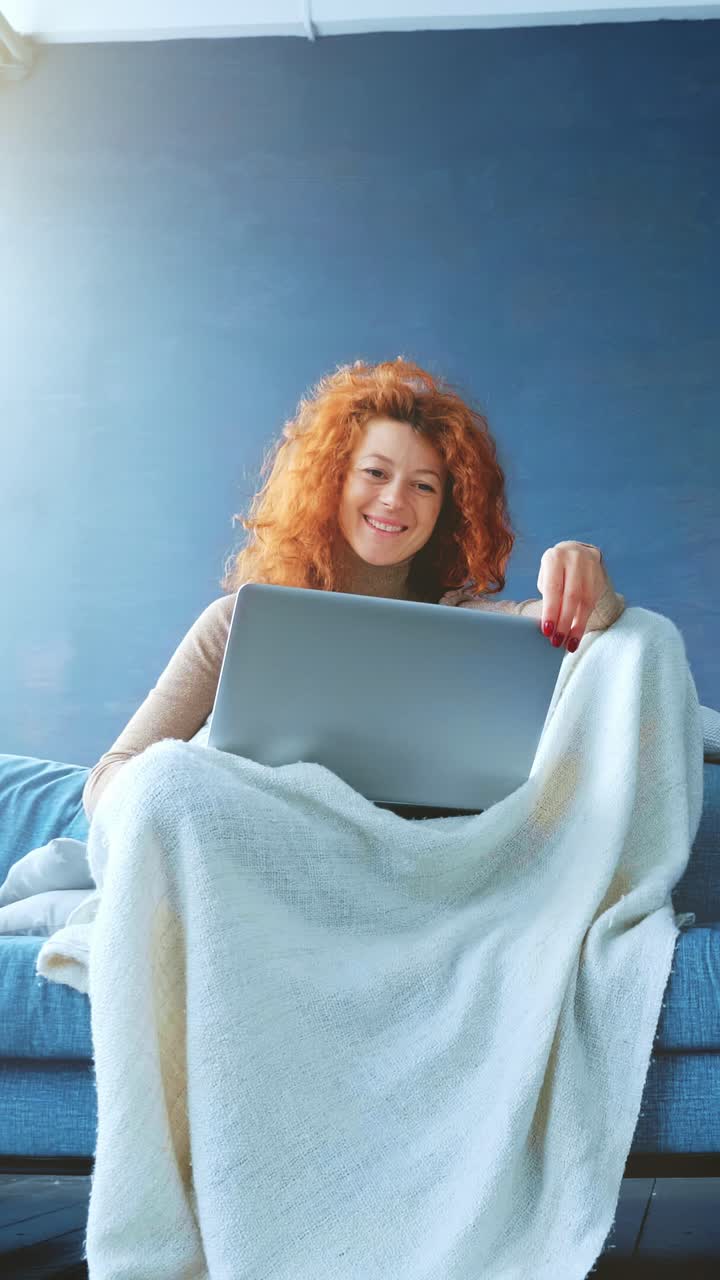 Young woman with curly red hair is sitting on a couch, wrapped in a cozy blanket, using a laptop while enjoying a relaxed indoor atmosphere