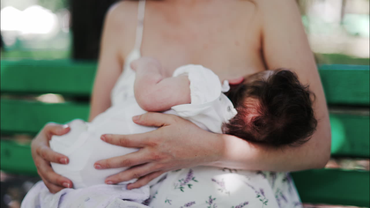 A mother siting on a bench in the park, breastfeeding her baby while enjoying a quiet, peaceful moment in nature