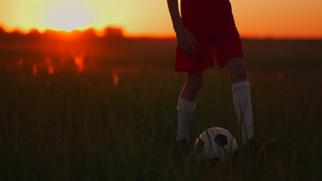 close-up of tracking the feet of a football boy in a red t-shirt and shorts running with the ball at sunset in the field on the grass. the young football player dreams of a professional career and trains in the field. on the way to success