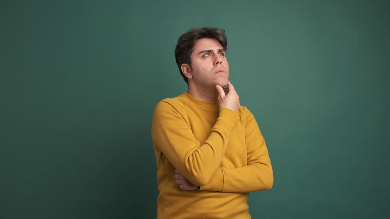 Thoughtful young man touching chin in green studio
