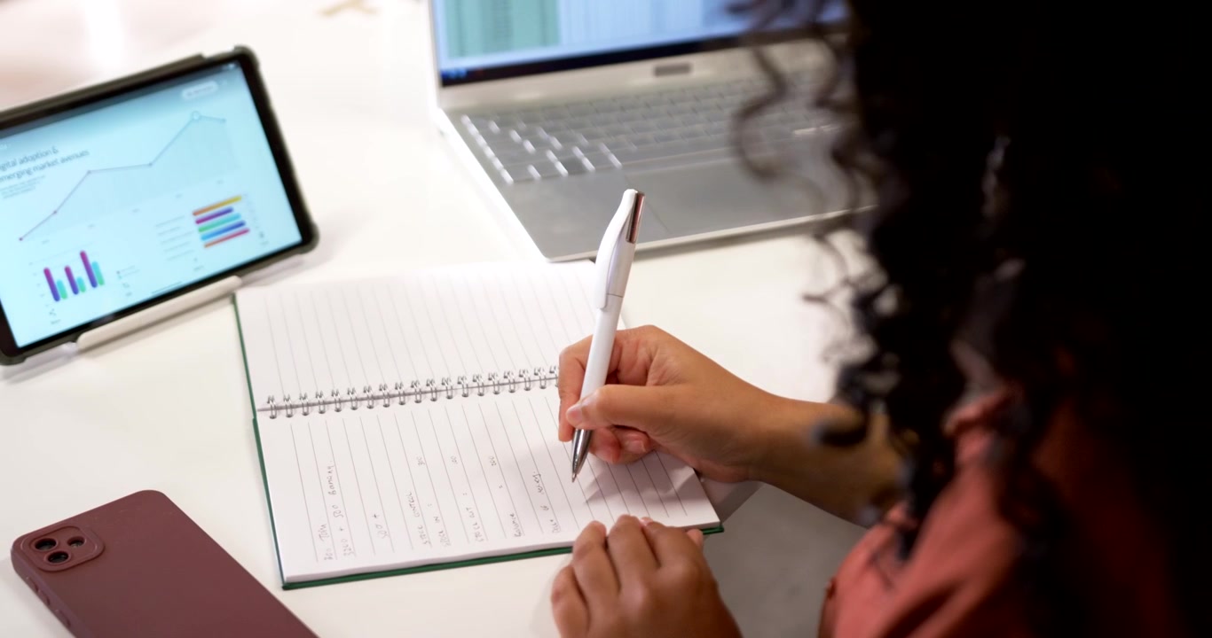 Pen is pointing at laptop spreadsheet with tablet charts on desk, notebook opening to record data