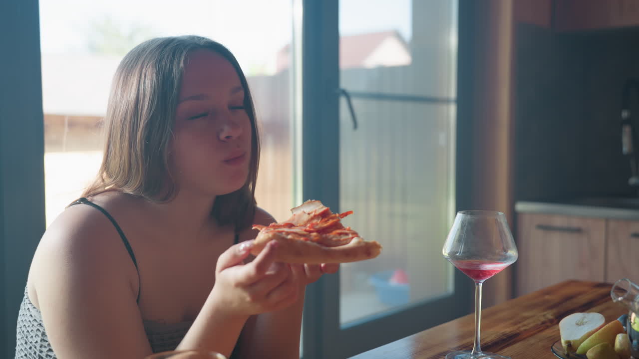 Elegant woman enjoying a slice of pizza with a glass of wine on a rustic table, captured in warm light, sharing good food with a friend in a cozy kitchen setting