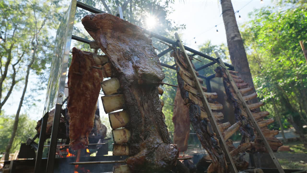 Large meat cuts including costillar, asado de tira, ribs and vacío hang vertically on iron frames, slowly roasting over embers during a traditional Argentine asado in a forest clearing under sunlight
