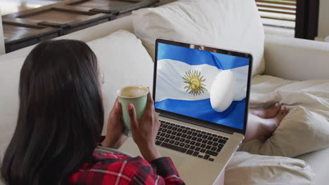 mujer biracial viendo una computadora portátil con una pelota de rugby con la bandera de argentina en la pantalla