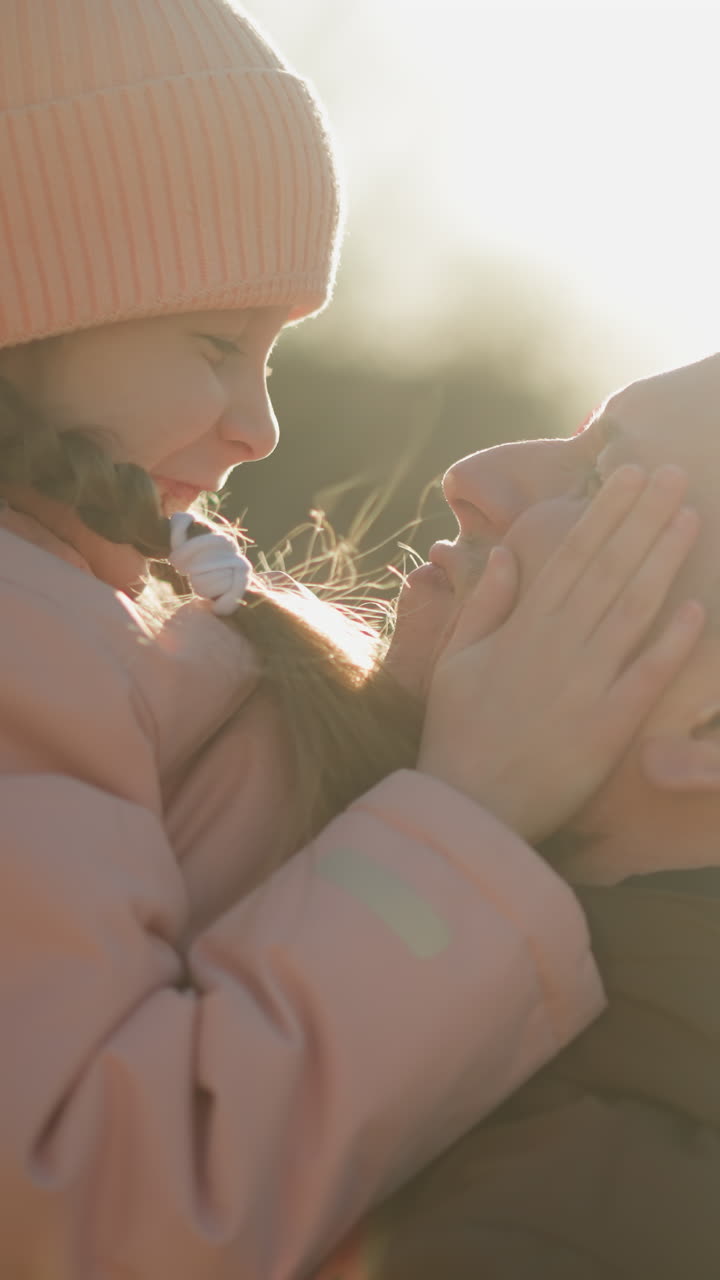핑크색 모자와 재을 입은 기쁜 어린 소녀가 갈색 재를 입은 남자에게 끌려가며, 그녀는 장난스럽게 그의 얼굴을 만지고 그를 단단히 포옹하고, 둘 다 따뜻한 미소를 지으며 부드러운 순간을 공유하고 있습니다.