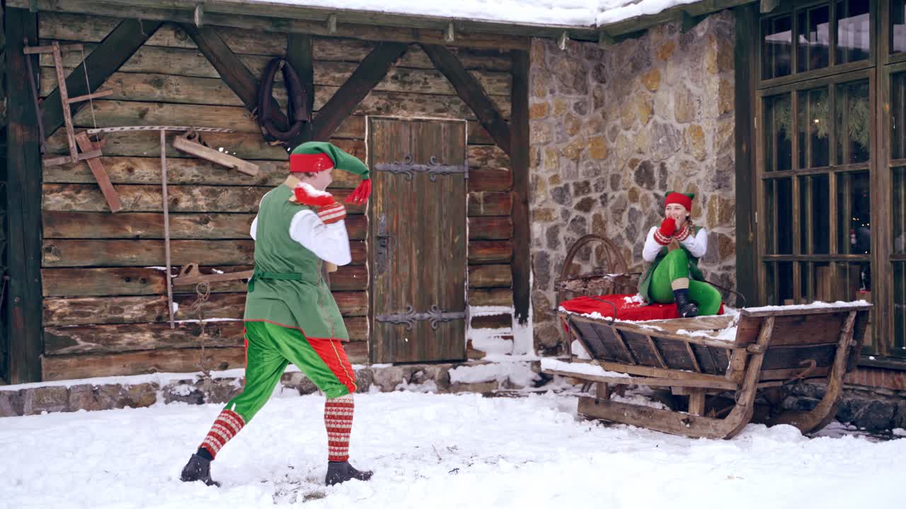 Joyful elves playing snowballs outdoors. Female elf sitting on wooden sleigh. Fairy elves in winter on the background of old wooden house. Christmas atmosphere.