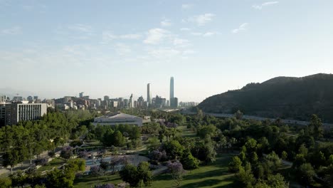 Vista-Aérea-Hacia-Atrás-Del-Parque-Bicentenario-De-Vitacura-En-La-Ciudad-De-Santiago-De-Chile-Con-El-Distrito-Financiero-En-El-Horizonte---Disparo-De-Drones