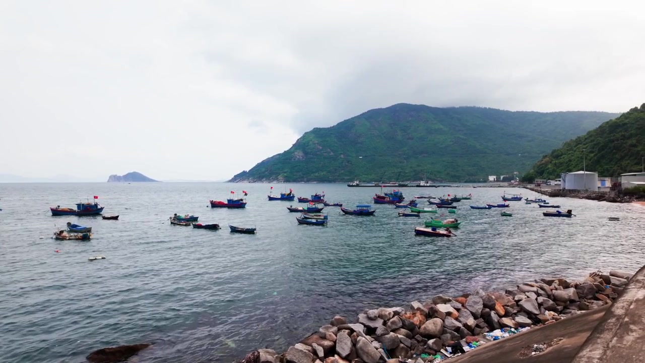 O porto em Dak Lak, Vietnã, mostra muitos barcos flutuando na água. O céu está nublado e montanhas se erguem ao longe. Pescadores e pessoas locais se movem pela área.