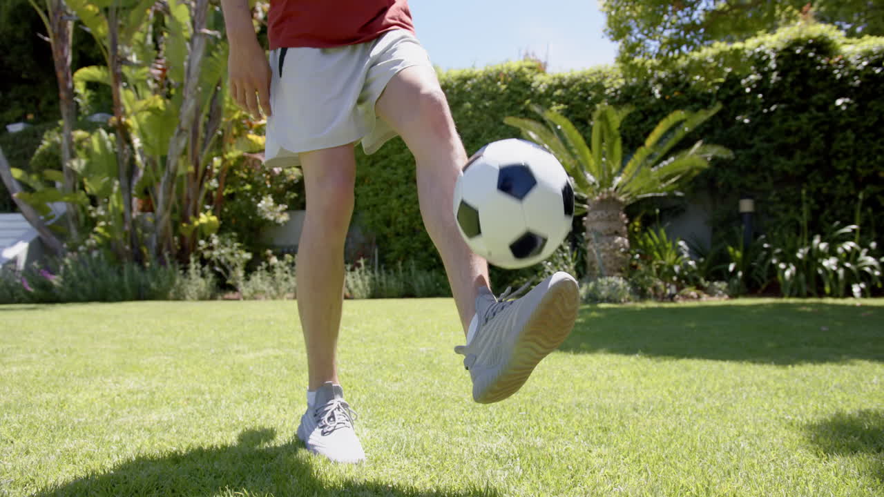 sección baja de hombre biracial malabarismo fútbol con los pies en el jardín soleado, copia espacio, cámara lenta