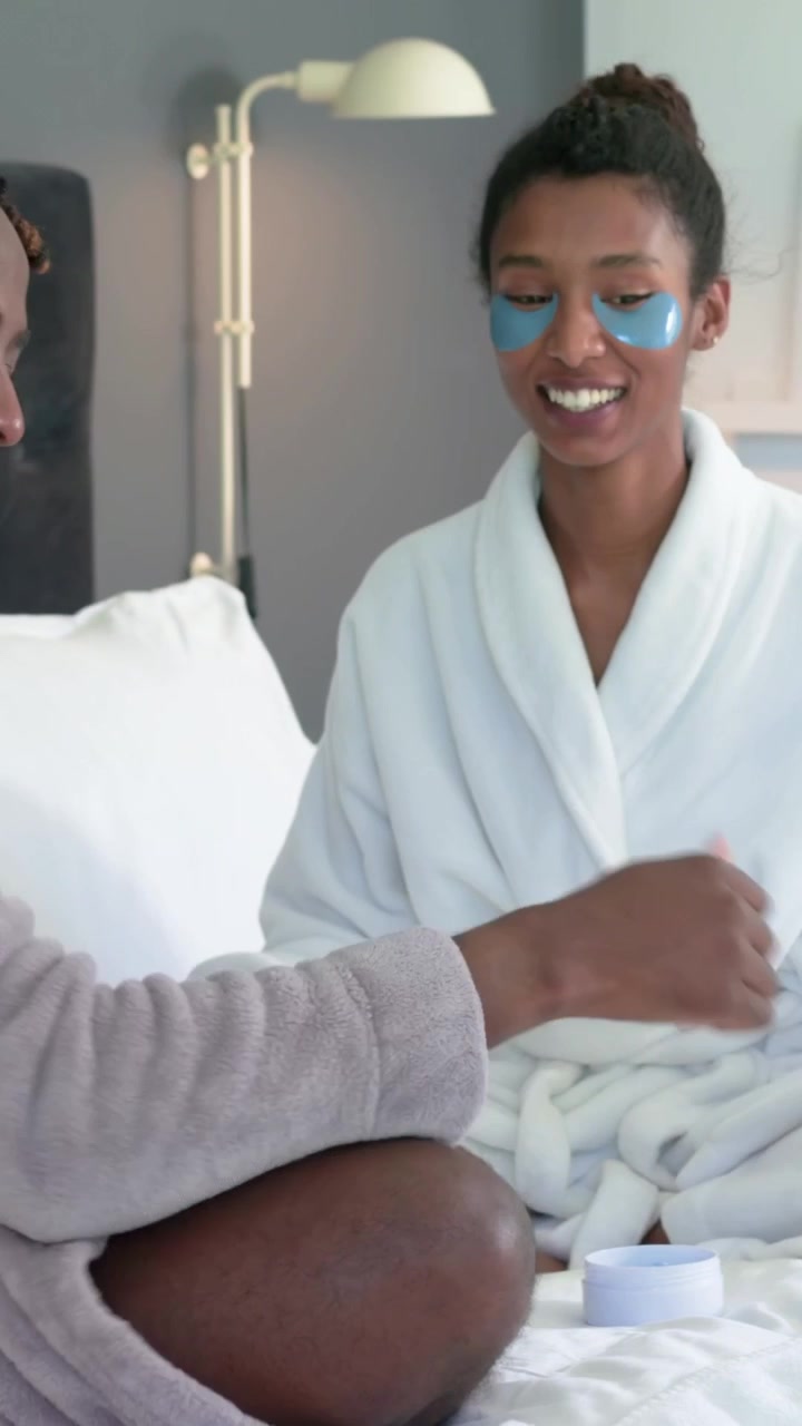 Vidéo verticale : Femme en peignoir tenant un miroir, laissant un homme lui appliquer des patchs pour les yeux sur le lit pour vérification.