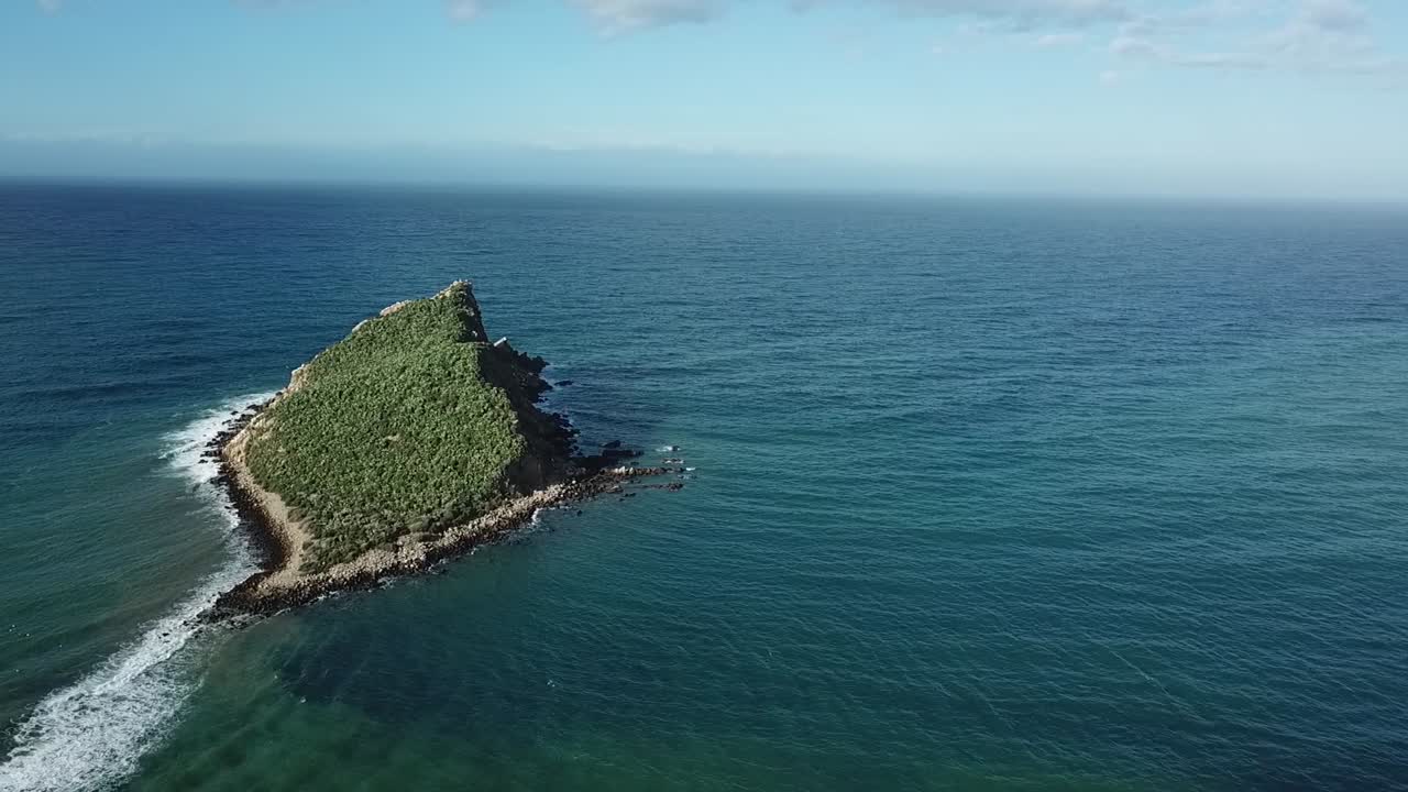 Aerial drone view of a small rocky islet located near a golden sandy beach. Clear turquoise Mediterranean waters surrounding the solitary rock formation