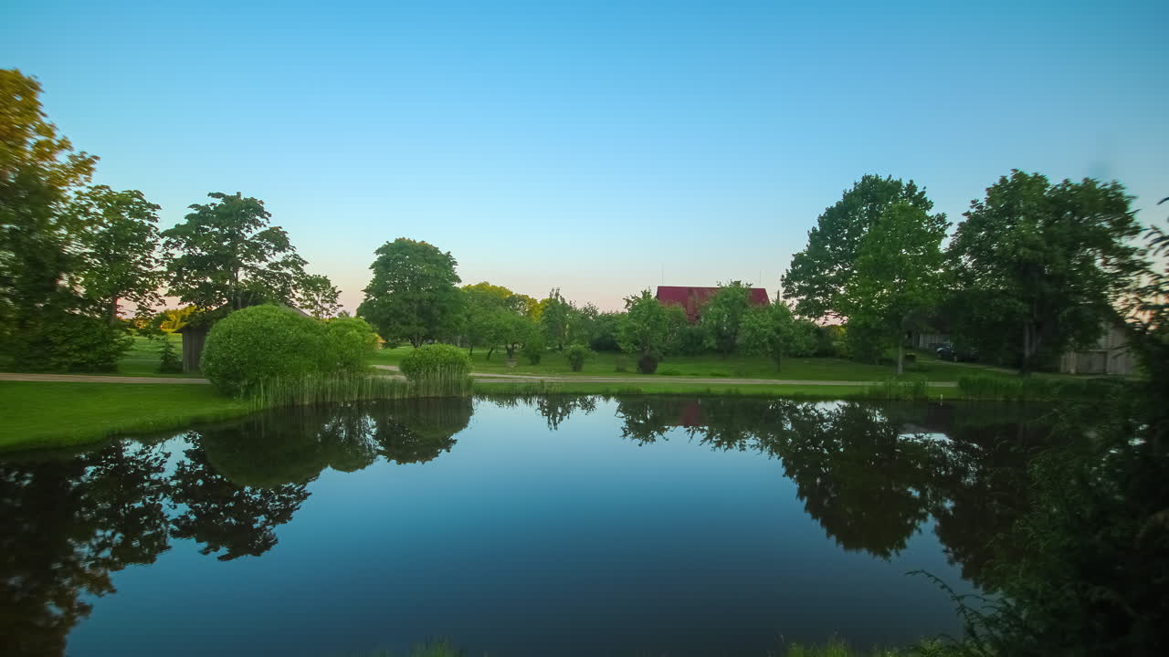 나무, 잔디, 뒷면에 빨간 지붕이 있는 집과 함께 평온한 반사 호수 위에 떠오르는 태양의 영화적 타임 스.