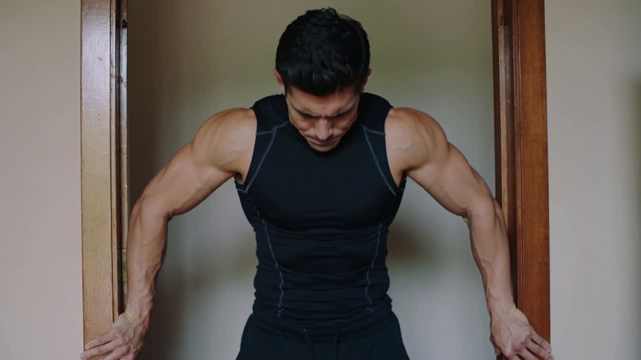 Gripping home jambs, man in black top performing shoulder shrugs for traps, dark rectangle right