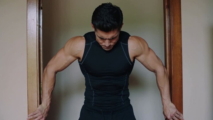 Gripping home jambs, man in black top performing shoulder shrugs for traps, dark rectangle right